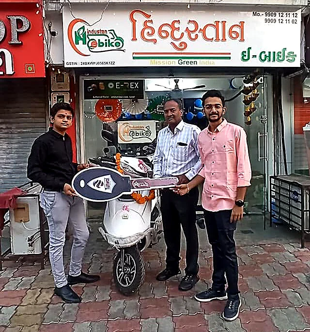 Customers collecting a new electric scooter outside the Hindustan E-Bike showroom in Nana Varachha, Surat, beneath the shop's Gujarati signboard